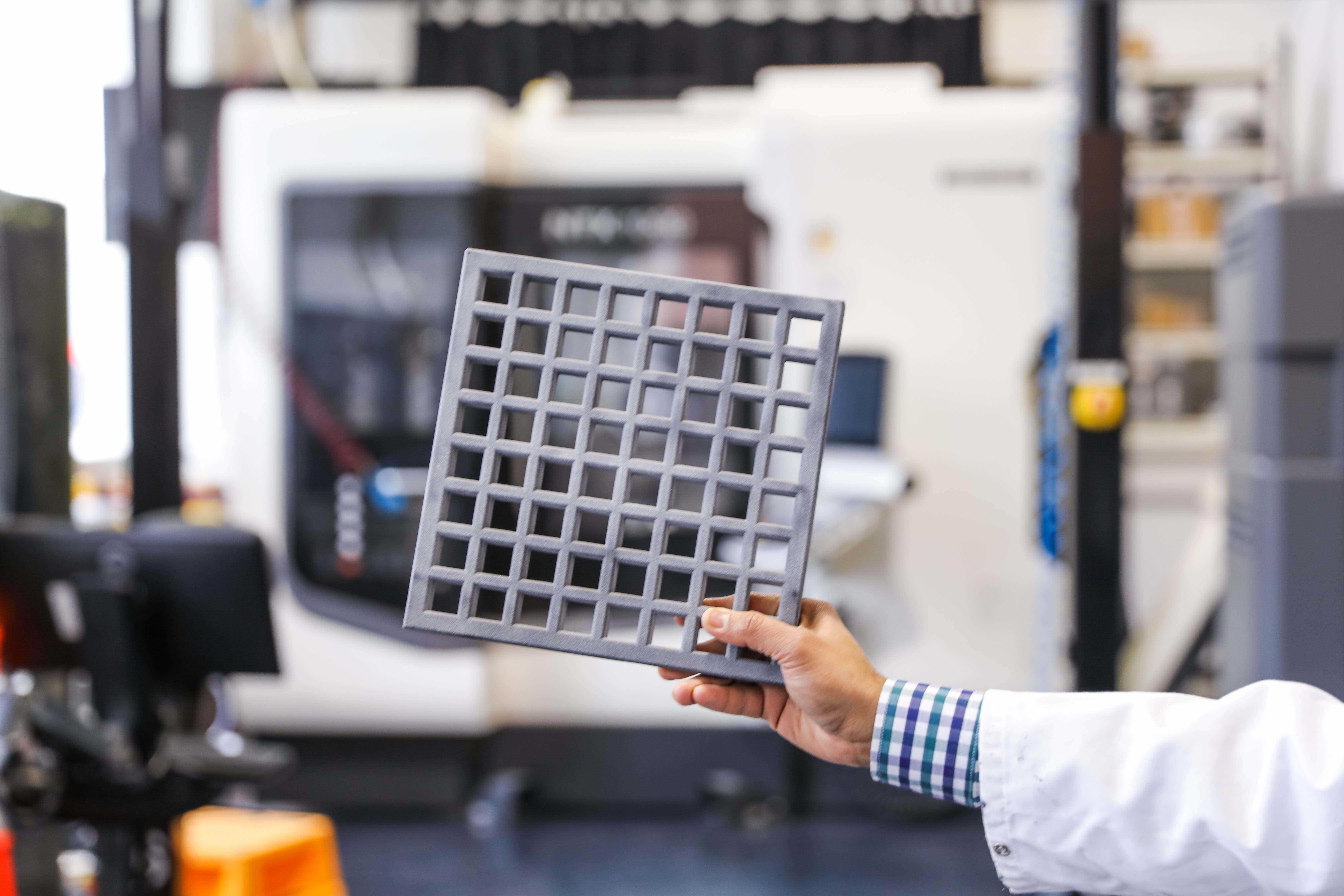 一名男子在实验室的3D打印机前拿着一个方形的剖面线3D打印灰色材料